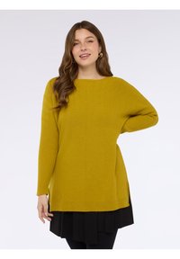Maglione a maniche lunghe giallo senape con texture a costine e spacchi laterali, caratterizzato da una scollatura rotonda e una vestibilità rilassata. Sotto, gonna nera a strati.