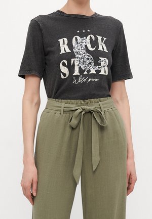 Person iført en sort "Rock Star Wild Power" t-shirt med en sølv paliet-kat og olivengrønne bukser med høj talje, bundet med et bælte med sløjfe.