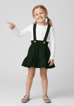 Uśmiechnięta dziewczynka o blond kucykach w białej koszulce z długim rękawem, czarnej sukience na szelkach i szarych butach wskazuje w lewo prawą ręką.