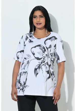 Woman with long black hair wearing a white shirt with large black and gray floral prints and black pants, standing against a white wall.