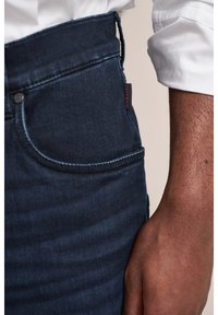 Ciemnoniebieskie dżinsy o dopasowanym kroju, z kontrastowym przeszyciem, przednią kieszenią i małą, czerwoną metką marki na pasku.