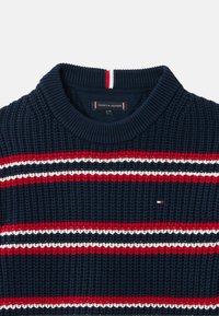 Marineblå strikkegenser med røde og hvite horisontale striper, ribbestrikket rund hals og liten logodetalj. Teksturert stoff med synlige sømmer.