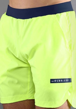 Pantalones cortos deportivos amarillos neón con cintura azul marino y logo "ENDLESS" en el muslo, usados por una persona.