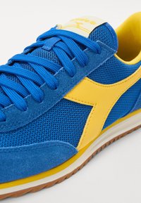 Blå sneakers med gul logotyp och detaljer, blå snörning, ovandel av mesh och mocka, vit och gul sula samt brun slitban.