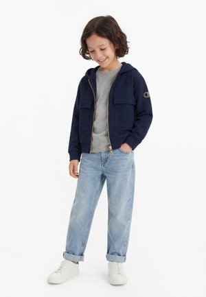 Kind met donker krullend haar glimlacht, draagt een marineblauwe zip-up hoodie, grijs T-shirt, lichtblauwe opgerolde spijkerbroek en witte sneakers.