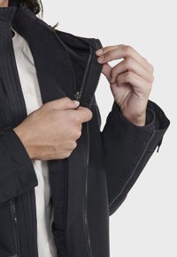 Veste noire zippée avec un col montant, présentant une texture lisse, des coutures contrastées et une doublure intérieure matelassée.