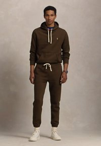 Brun bomull hettegenser og matchende joggebukser med snøring. Hvite detaljer og logo på brystet. Brukt med hvite sneakers.