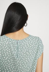 Femme aux longs cheveux noirs vue de dos, portant un haut vert à pois avec une fermeture éclair dans le dos et une boucle d’oreille argentée torsadée en forme d’anneau.