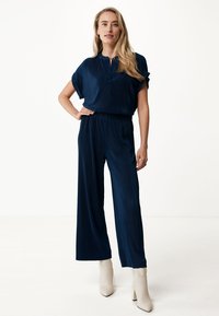 Marinblå, plisserad jumpsuit med korta ärmar, framplacket och elastiskt midjeband, matchad med krämfärgade ankelstövlar.