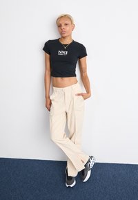 Svart kortärmad t-shirt med Nike-logotyp, beige skräddarsydda byxor med sidofickor och svarta sneakers med vita detaljer. Ljust bakgrund.