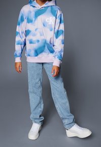 Person som bär en blå och vit tie-dye hoodie med "cK"-logotyp, ljusblå jeans och vita sneakers på en grå bakgrund.