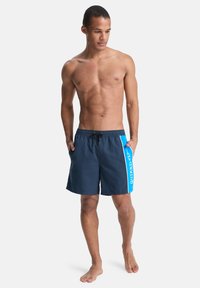 Μπλε σκούρο μαγιό σορτς με μπλε διακοσμητικό πάνελ, με λογότυπο, ελαστική μέση με κορδόνι και πλαϊνές τσέπες. Λείο ύφασμα.