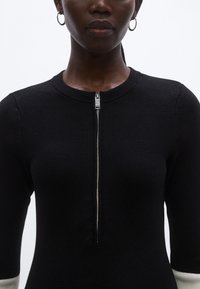 Čierne priliehavé svetrové šaty s okrúhlym výstrihom a krátkymi rukávmi. Majú predný zips a kontrastné biele manžety. Textúrovaná pletenina.