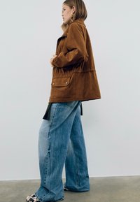 Veste en daim marron avec une coupe décontractée, poches latérales et coutures visibles, associée à un jean large bleu clair et des chaussures à motifs.