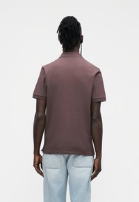 Braunes Polohemd mit kurzen Ärmeln aus Baumwolle, mit kleinem Kragen und geradem Saum. Getragen zu hellblauen Jeans aus Denim.