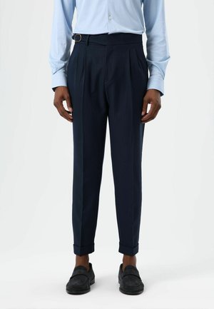 Marineblaue plissierte Hose mit einem Bundverschluss, schmal geschnitten und mit umgeschlagenen Säumen, aus strukturiertem Stoff. Kombiniert mit dunklen Loafers.