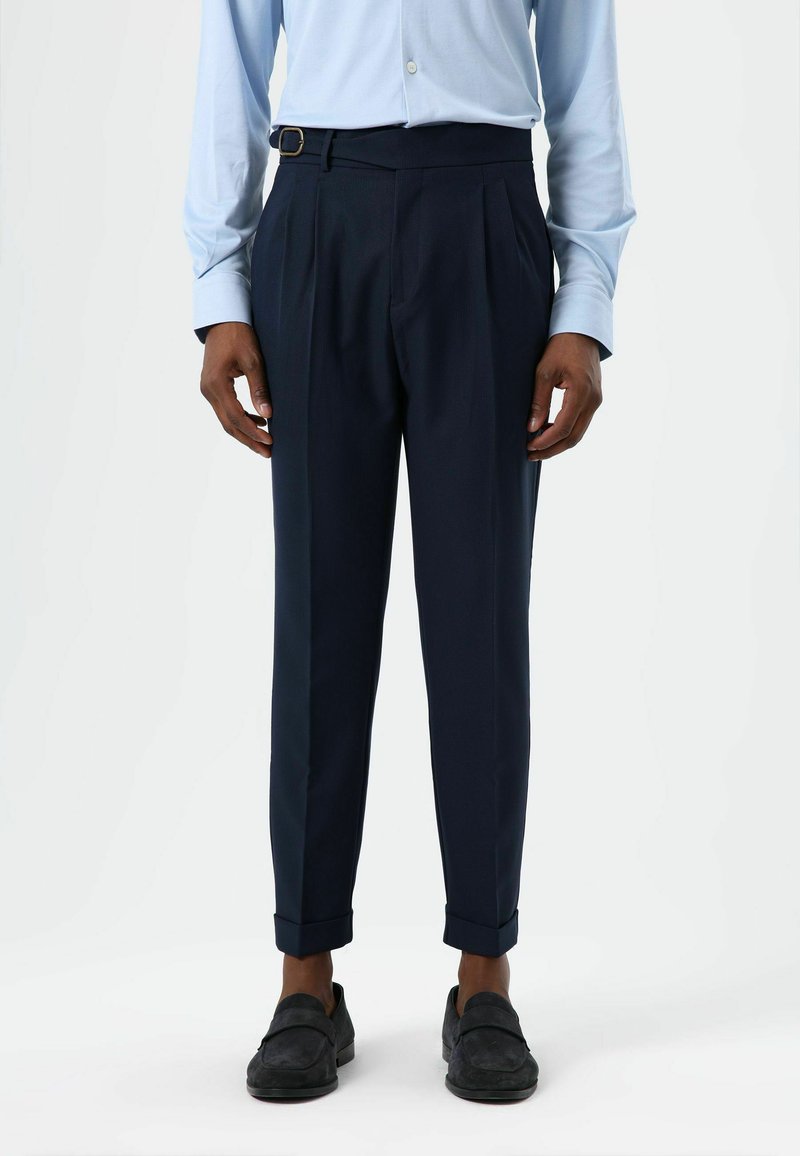 Marineblauwe geplooide broek met een taillebandgesp, voorzien van een getailleerde pasvorm en omslagboorden, gemaakt van textuurstof. Draagt donkere loafers.