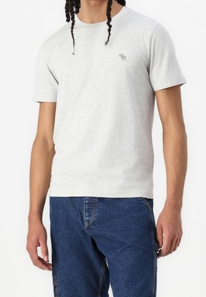 Mann trägt hellgraues kurzärmeliges T-Shirt mit kleinem Elch-Logo auf der linken Brust und lockere blaue Jeans.