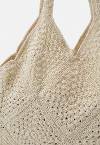 Vaaleanbeige virkattu laukku, jossa on teksturoitu timanttikuvio ja kaksi leveää kantohihnaa, esittelee käsintehtyä suunnittelua.