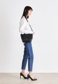 Svart läderhandväska med sidohandtag, slät textur, rektangulär form, buren över axeln av en person i vit skjorta och blå jeans.