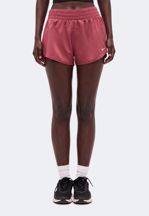 Persona che indossa pantaloncini sportivi bordeaux, sneakers nere con suola bianca, calzini bianchi e un crop top nero, in piedi su uno sfondo semplice.