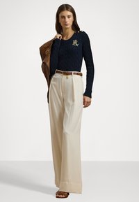 Maglione blu navy a trecce con dettagli in metallo dorato, abbinato a pantaloni beige a gamba larga e giacca di pelle marrone. Cintura marrone con charm dorato.