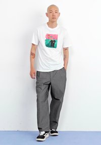 Hvid T-shirt med et farverigt grafikdesign, der viser blomster og en figur; samt med grå bukser og sorte sneakers med hvide såler.