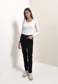 Valkoinen pitkähihainen paita, mustat korkeavyötäröiset farkut edustataskuilla, yhdistetty beigen ja valkoisen värisiin lenkkareihin, vaaleaa taustaa vasten.