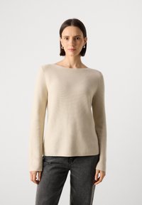 Suéter bege de manga longa com decote barco, feito de um material texturizado, emparelhado com jeans cinza-escuro. Design simples e ajustado.