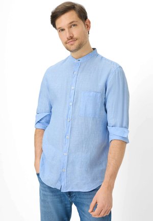 Mann trägt hellblaues Hemd mit hochgekrempelten Ärmeln und blaue Jeans, steht mit den Händen in den Taschen vor weißem Hintergrund.