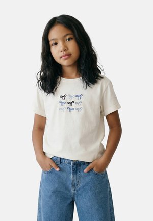 Fille aux cheveux noirs ondulés portant un t-shirt blanc avec des nœuds brodés bleus et noirs, et un jean en denim bleu, mains dans les poches.