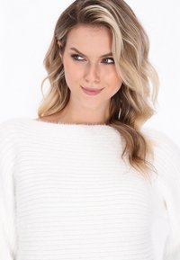 White striped sweater with a loose neckline, soft texture, and long sleeves. Model has wavy hair and is looking slightly to the side.