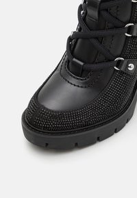 Stivale alla caviglia nero con una tomaia in pelle liscia e un pannello di strass testurizzato. Design con lacci e dettagli metallici, suola spessa.