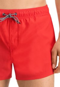 Rote Bade Shorts aus einem leichten Stoff, ausgestattet mit einem elastischen Bund und einem Kordelzug sowie einer kleinen Seitentasche.