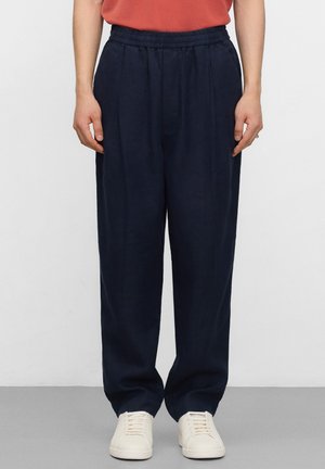 Osoba nosząca granatowe, luźne spodnie z elastycznym pasem i białe trampki, stojąca na tle jednolitego jasnego tła.