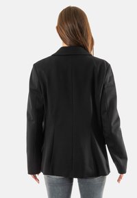 Blazer nero su misura con spalline strutturate, rever accentuati e silhouette aderente. Realizzato in tessuto liscio e intrecciato con tasche laterali.