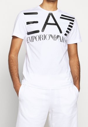 Mann trägt weißes Emporio Armani EA7 T-Shirt mit schwarzem, fettem Logo und weiße Hose, Hände in den Taschen, Bildausschnitt auf Hals und Oberschenkel.