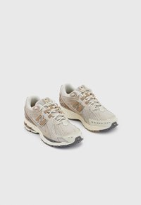 Chaussures de course en mesh beige et crème avec des accents gris et beiges. Elles comportent des lacets, une semelle coussinée et une surface extérieure texturée.