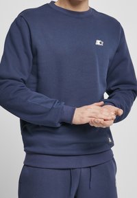 Persona vistiendo una sudadera azul marino y pantalones de chándal a juego, con pequeños logos de estrellas blancas en el pecho y en la cintura.
