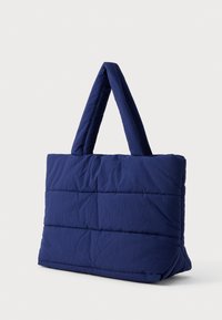 Námorně modrá polstrovaná taška s širokými uchy, obdélníkový tvar a vodorovné švy na čistě bílém pozadí.
