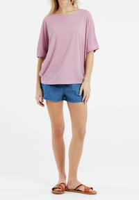 Nainen, joka käyttää löysää, vaaleanpunaista siksakkuvioista yläosaa, sinisiä farkkushortseja ja ruskeita liukusandaaleja, seisoo valkoista taustaa vasten.