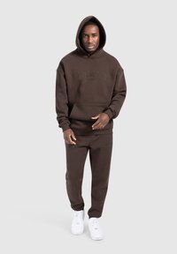Sudadera marrón con un bolsillo frontal y un logo bordado, combinada con pantalones de chándal a juego. Zapatillas blancas completan el conjunto.