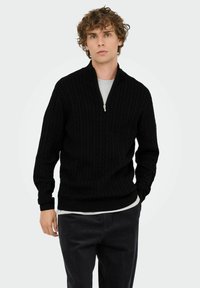 Junger Mann mit lockigem Haar, der einen schwarzen Strickpullover mit halbem Reißverschluss über einem weißen Hemd und einer dunklen Hose trägt, vor einem schlichten Hintergrund.