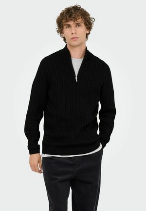 Junger Mann mit lockigem Haar, der einen schwarzen Strickpullover mit halbem Reißverschluss über einem weißen Hemd und einer dunklen Hose trägt, vor einem schlichten Hintergrund.