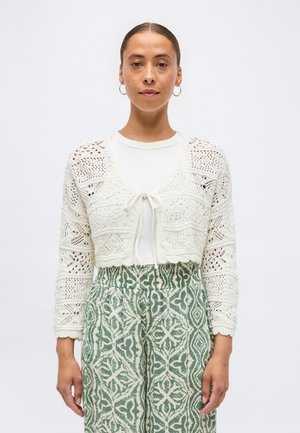 Naine, kes kannab valget kootud lühikest kardigani, mis on ees seotud, valget toppi ja kõrge vöökohaga rohelist mustrilist pükst, seisab tavalise taustaga ees.