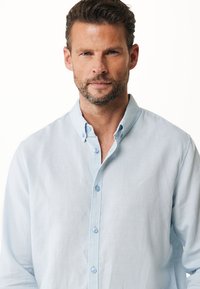 Camisa de botón de manga larga en azul claro, hecha de tela de algodón. Presenta un cuello clásico y botones en el frente. Textura suave, sin patrones.