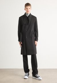 Černý dvouřadý trenčkot s límcem, kombinovaný s černými kalhotami a černo-bílými teniskami, stojící proti jednobarevnému pozadí.