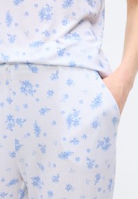 Σετ λευκών πιτζάμων με μικρό μπλε ανθισμένο μοτίβο, χέρι στην δεξιά τσέπη, ορατή υφή μαλακού, ελαφριού υφάσματος.