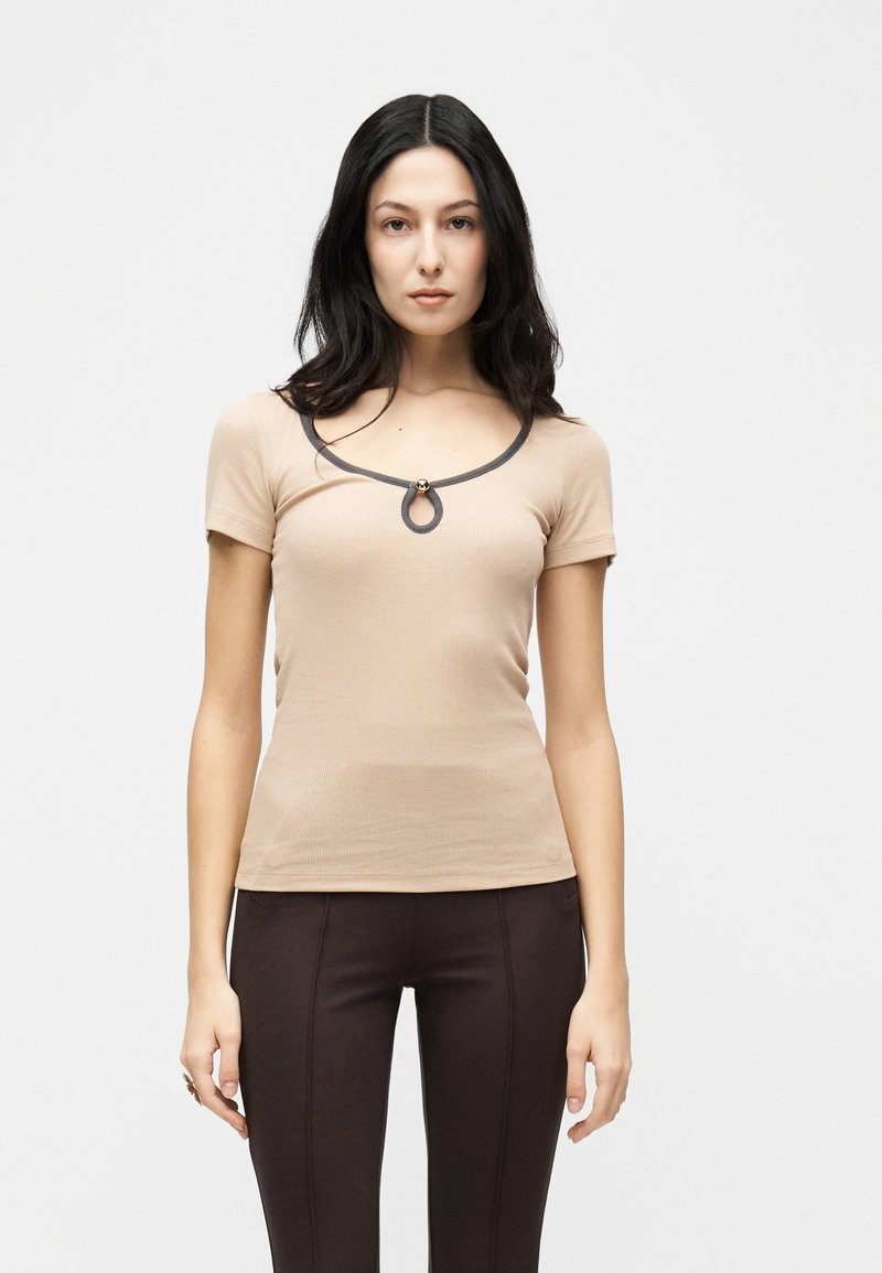 Γυναίκα με μακριά σκούρα μαλλιά που φοράει μπεζ κοντομάνικη μπλούζα με άνοιγμα στο ντεκολτέ και καφέ σκούρο στενά παντελόνια, στέκεται μπροστά σε λευκό φόντο.