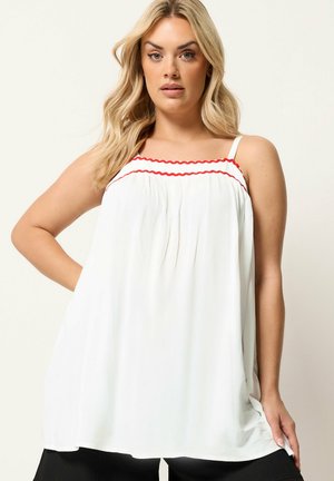 Blondynka w białej bluzce bez rękawów z czerwonym falistym paskiem na wysokości piersi, stoi z jedną ręką na biodrze.